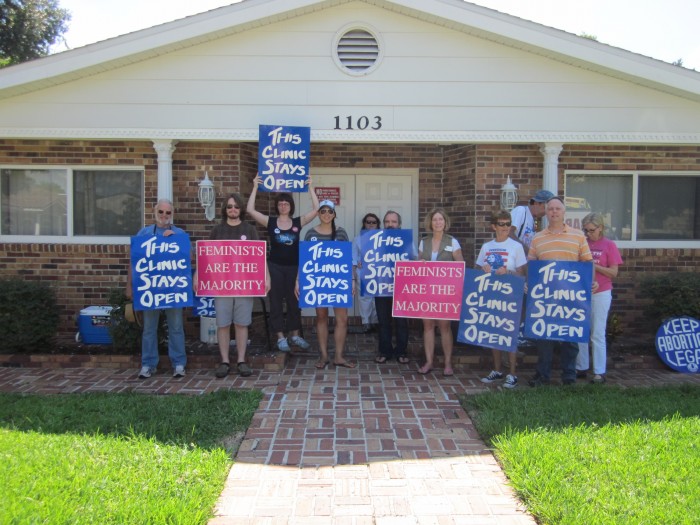 Supporters stand outside of a clinic with "This Clinic Stays Open" and "Feminists are the Majority" signs