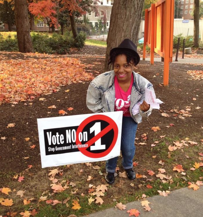 Feminist student wearing hat and denim jacket crouches next to a yard sign that reads "Vote NO on 1 Stop Government Interference" while on holding a stack of flyers in their hand and under their arm. Fallen leaves on the campus indicate it is fall.